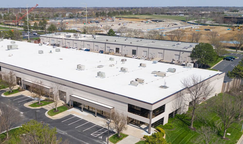 commercial-building-overhead Aerial view of a large white-roofed industrial building with parking spaces. Surroundings include trees, a paved road, and a construction site in the background.