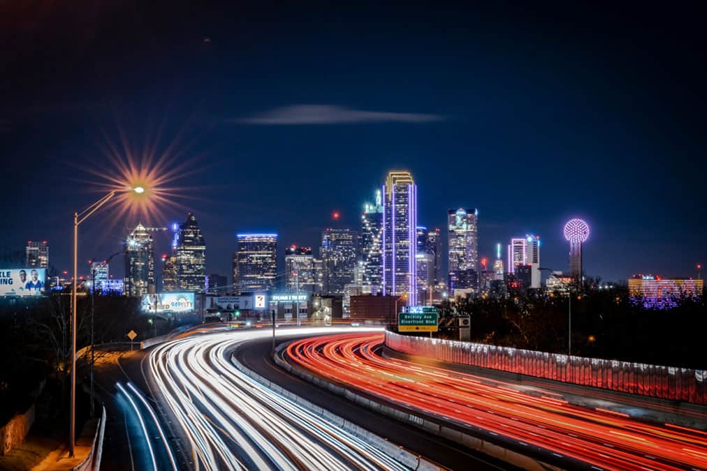 dallas-skyline-night Night view of a Dallas city skyline with brightly lit skyscrapers, including Reunion Tower, and light trails from vehicles on a highway in the foreground.