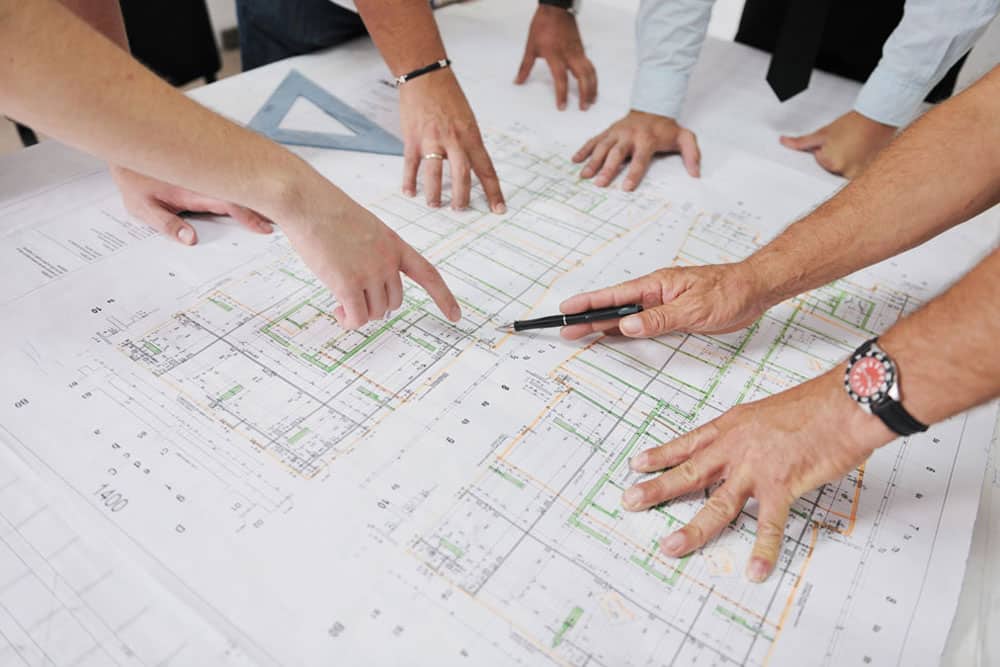 team-architects-construction-blueprints Several hands pointing at and gesturing over architectural blueprints spread out on a table.